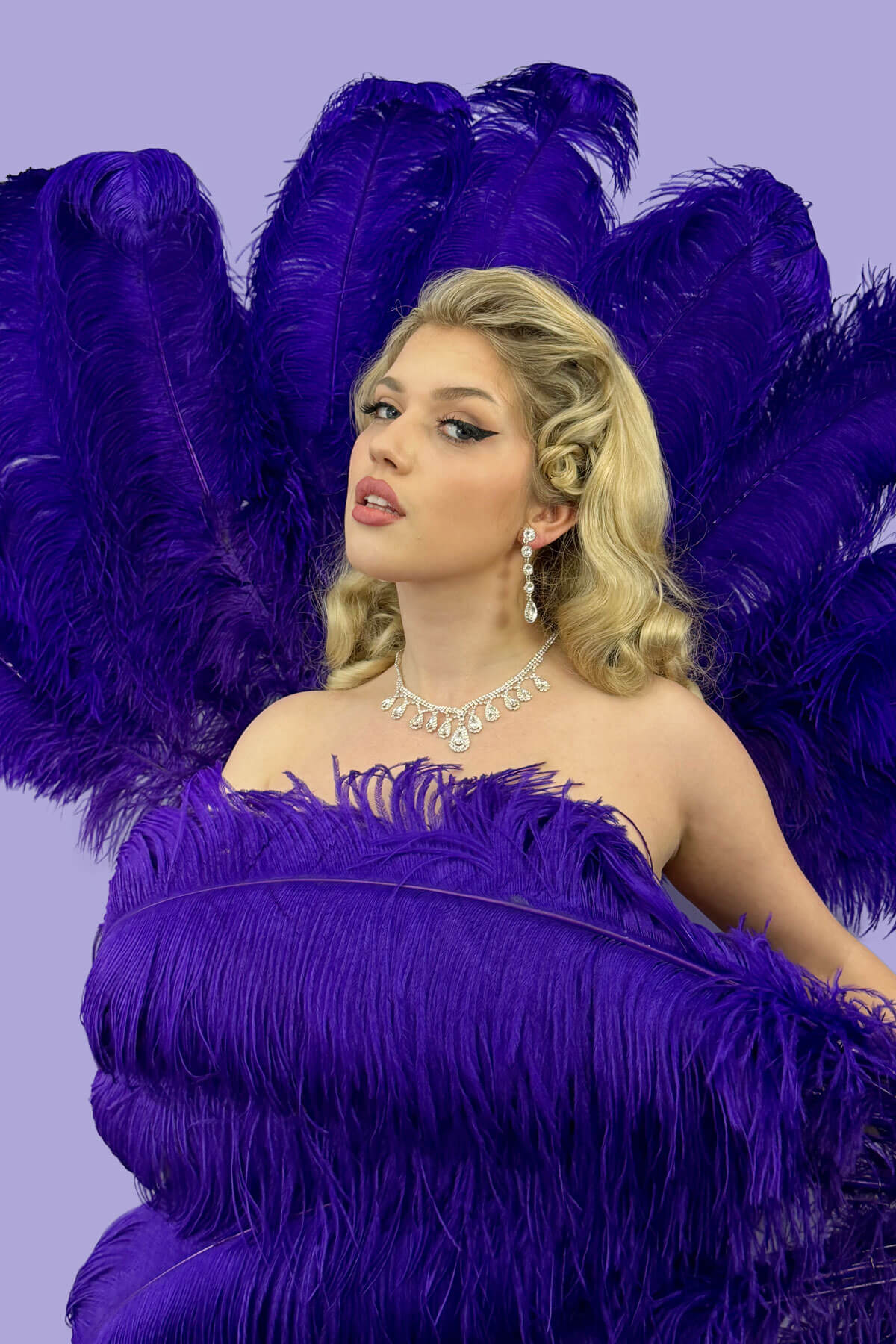 Woman in a purple feathered dress with a matching feathered headpiece against a purple background. 1950s style half wig hairpiece with deep finger waves: Deborah blonde 613T24