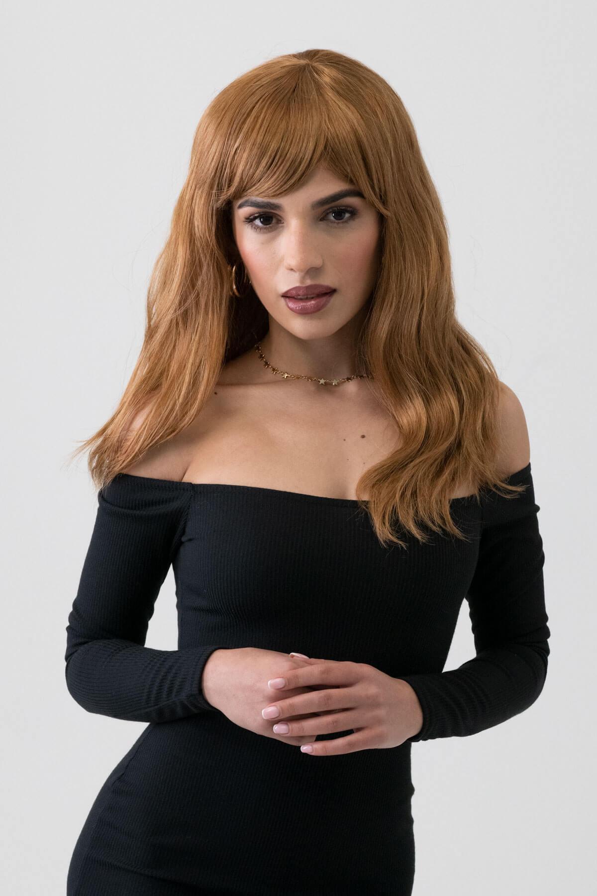 Woman with long brown hair wearing a black off-shoulder top against a white background. Golden brown wig with long waves: Carmella