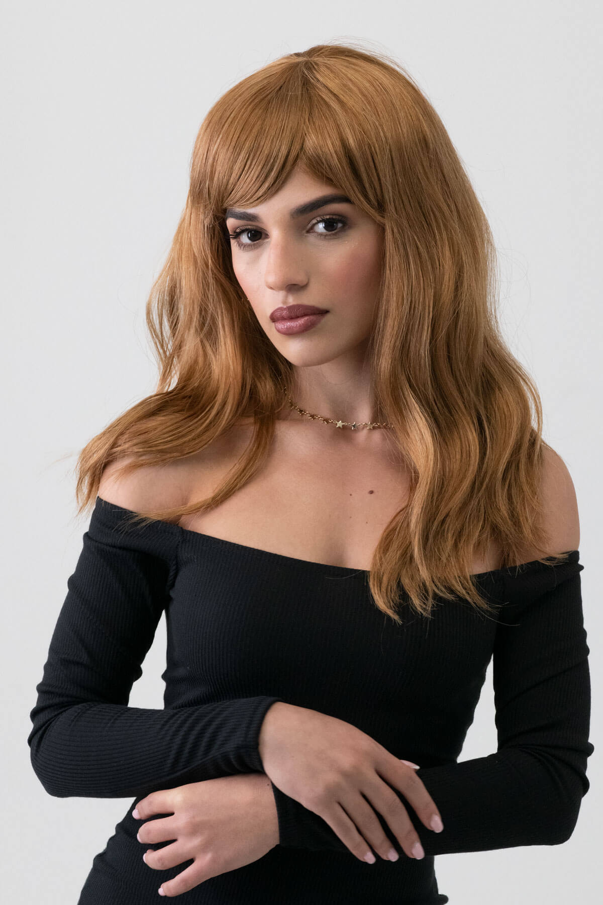 Woman with long brown hair wearing a black off-shoulder top against a white background. Golden brown wig with long waves: Carmella