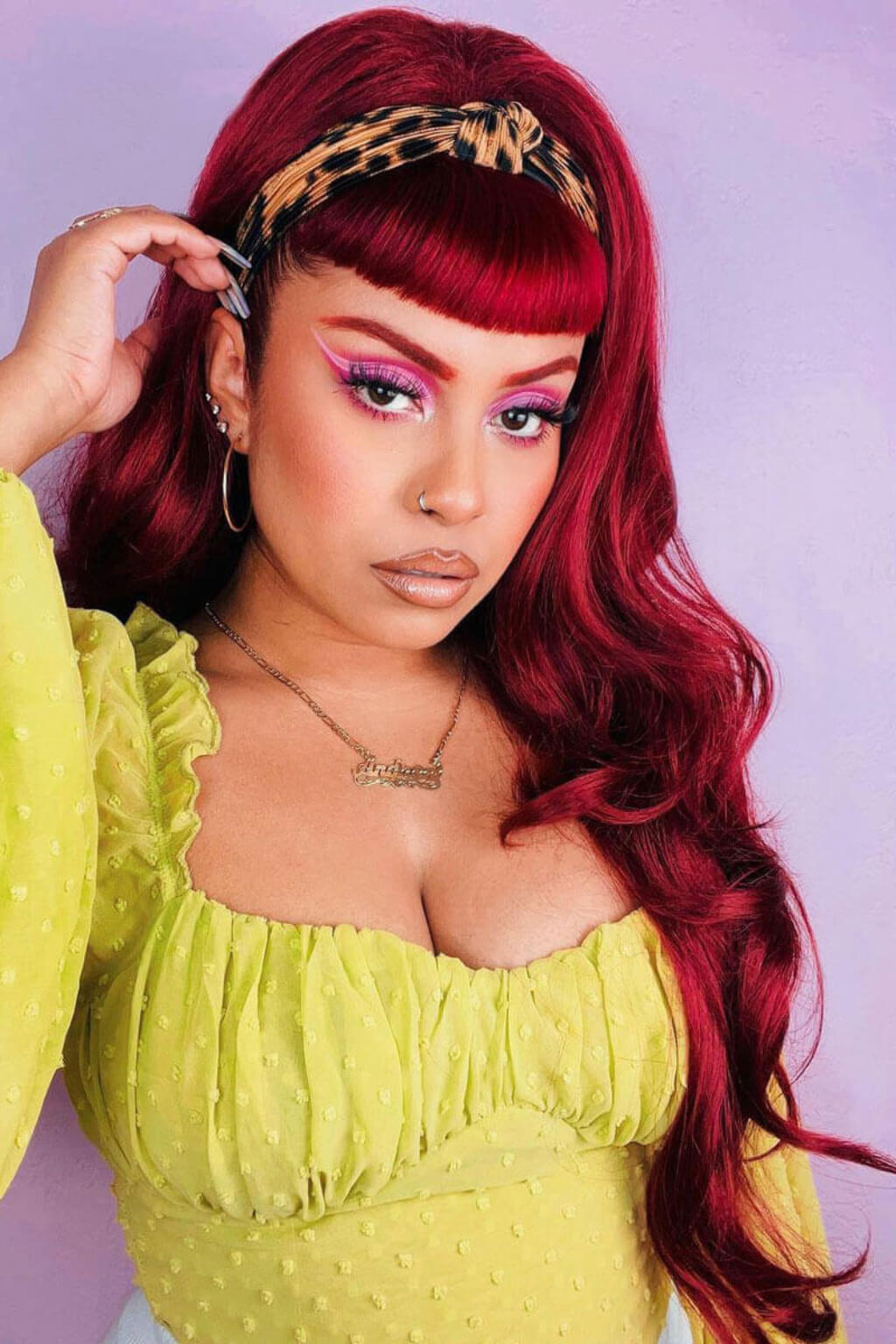 Woman with vibrant red hair and colourful makeup wearing a yellow top against a light purple background. High crown long, wavy half wig and volume ring: Bardot