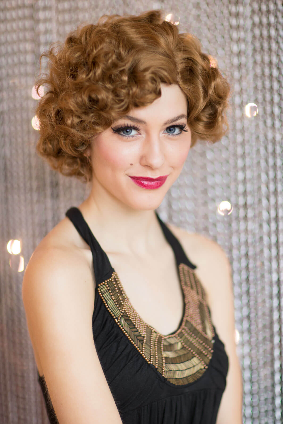 Woman with styled hair wearing a black dress with gold detailing against a textured silver background. Brown 1920s 1930s style wig, short retro style with finger waves: Cecelia light brown 12