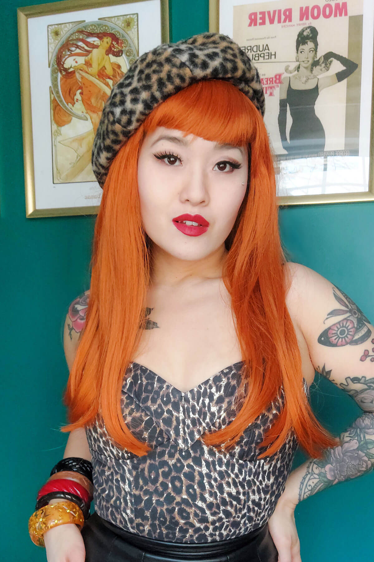 Person with orange hair and leopard print headband in front of a teal wall with framed posters. Long ginger wig with straight hair and a short, straight fringe: Daphne