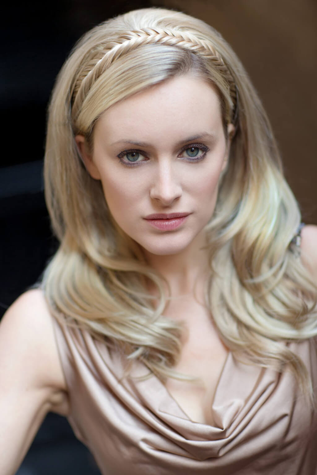 A close-up portrait of a blonde woman wearing a metallic champagne-brown draped top and a matching braided headband, set against a dark, softly blurred background. Fishtail plait hairband (braid) blonde