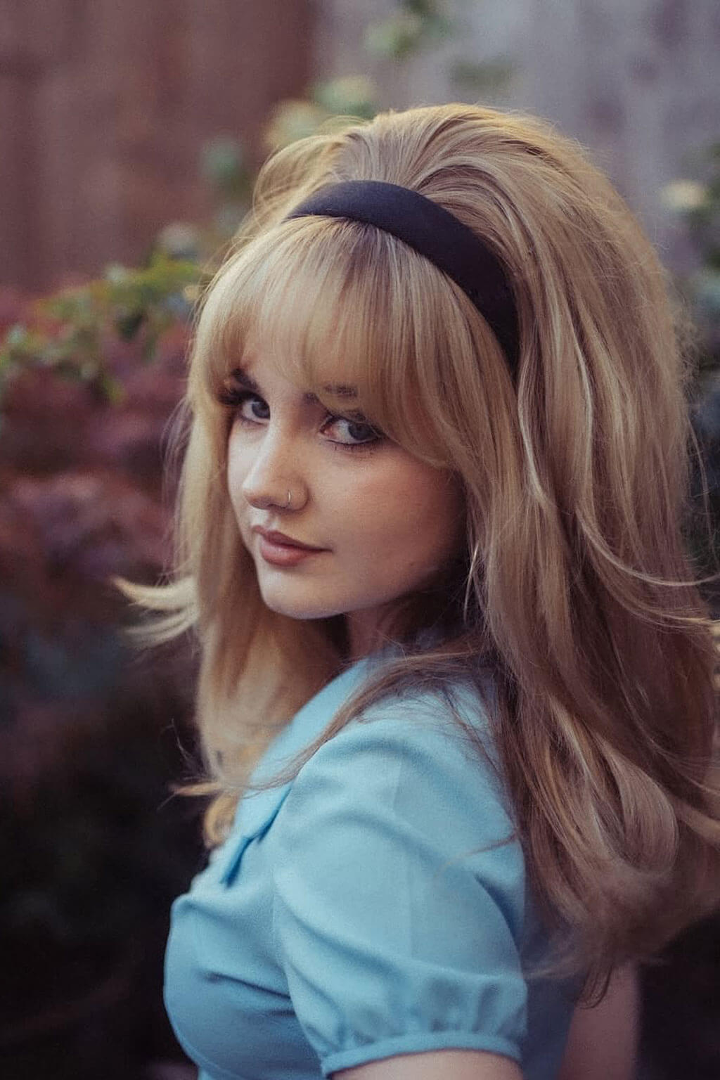 Woman with blonde hair and a blue top in an outdoor setting. Half wig hairpiece, long and wavy, Flexihair (Heat styleable) and volume ring for high crown: Sharon Blonde 24H613