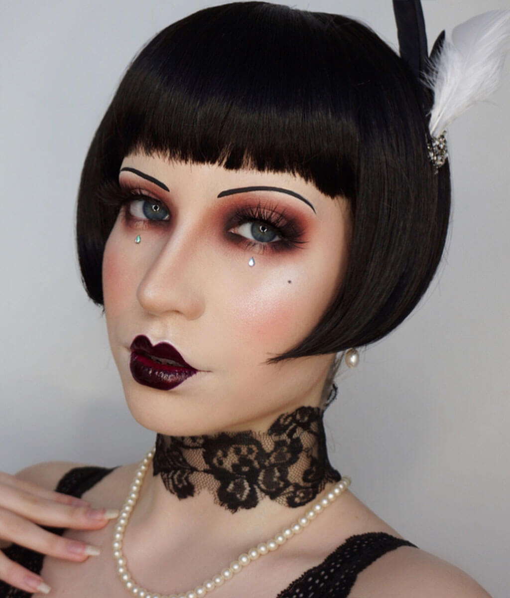 Person with vintage-style makeup and black bob wig with feather accessory on gray background. Louise Brooks style wig, short black bob: Brooks