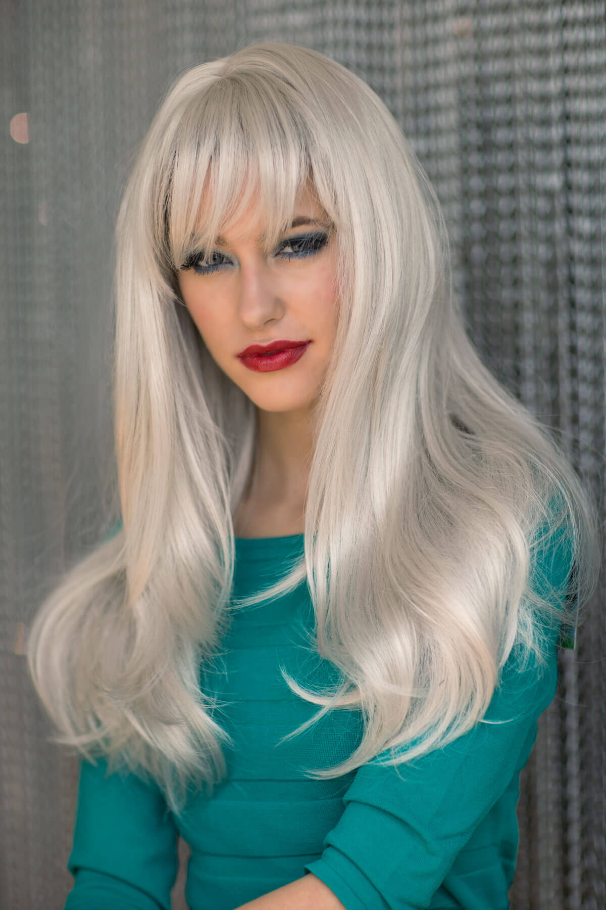 Person with long blonde wig and teal top against a textured grey background. Long silver/grey wig in razor cut, face framing style: London