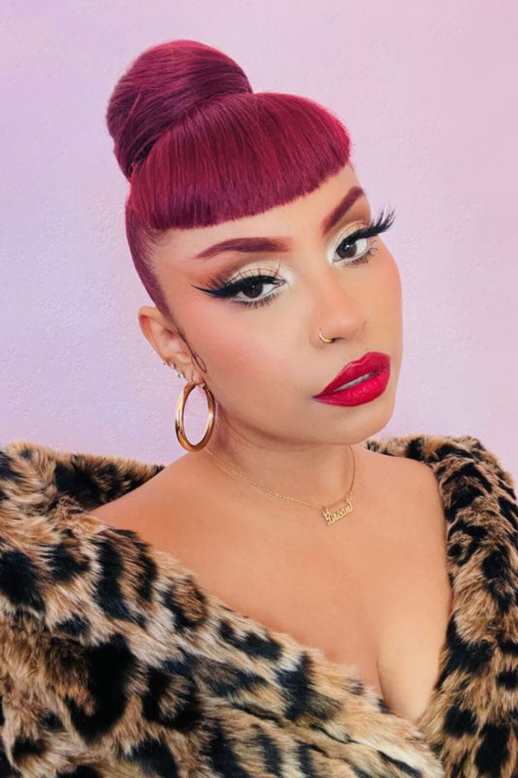Person with vibrant pink hair styled in an updo, wearing a leopard print garment against a light purple background. Hairpiece bun; instant perfect bun hairpiece, cherry red: Hepburn