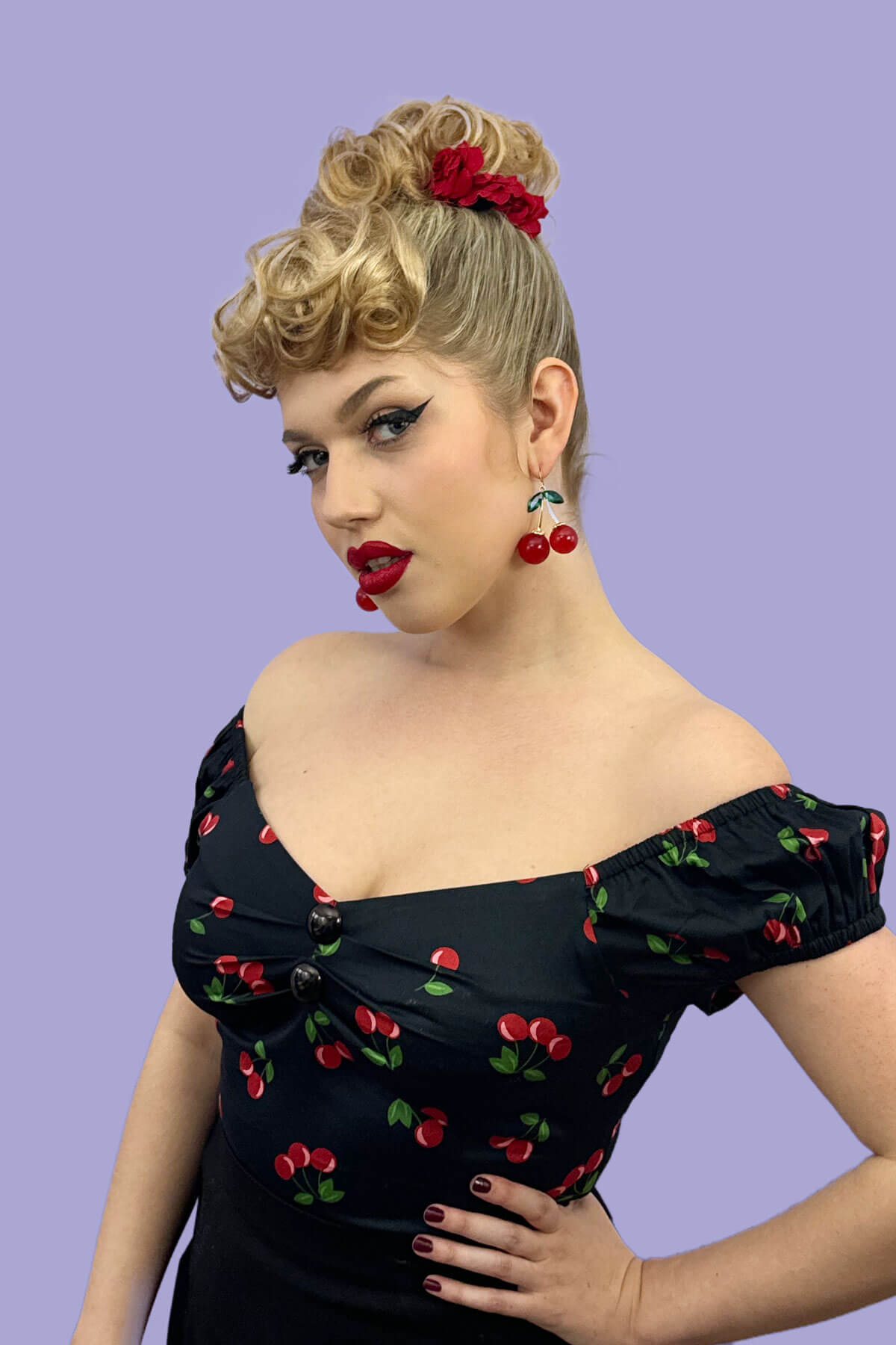 Woman wearing a black dress with red cherry patterns against a purple background. Curly bun hairpiece, rockabilly style: Toni blonde 613T24