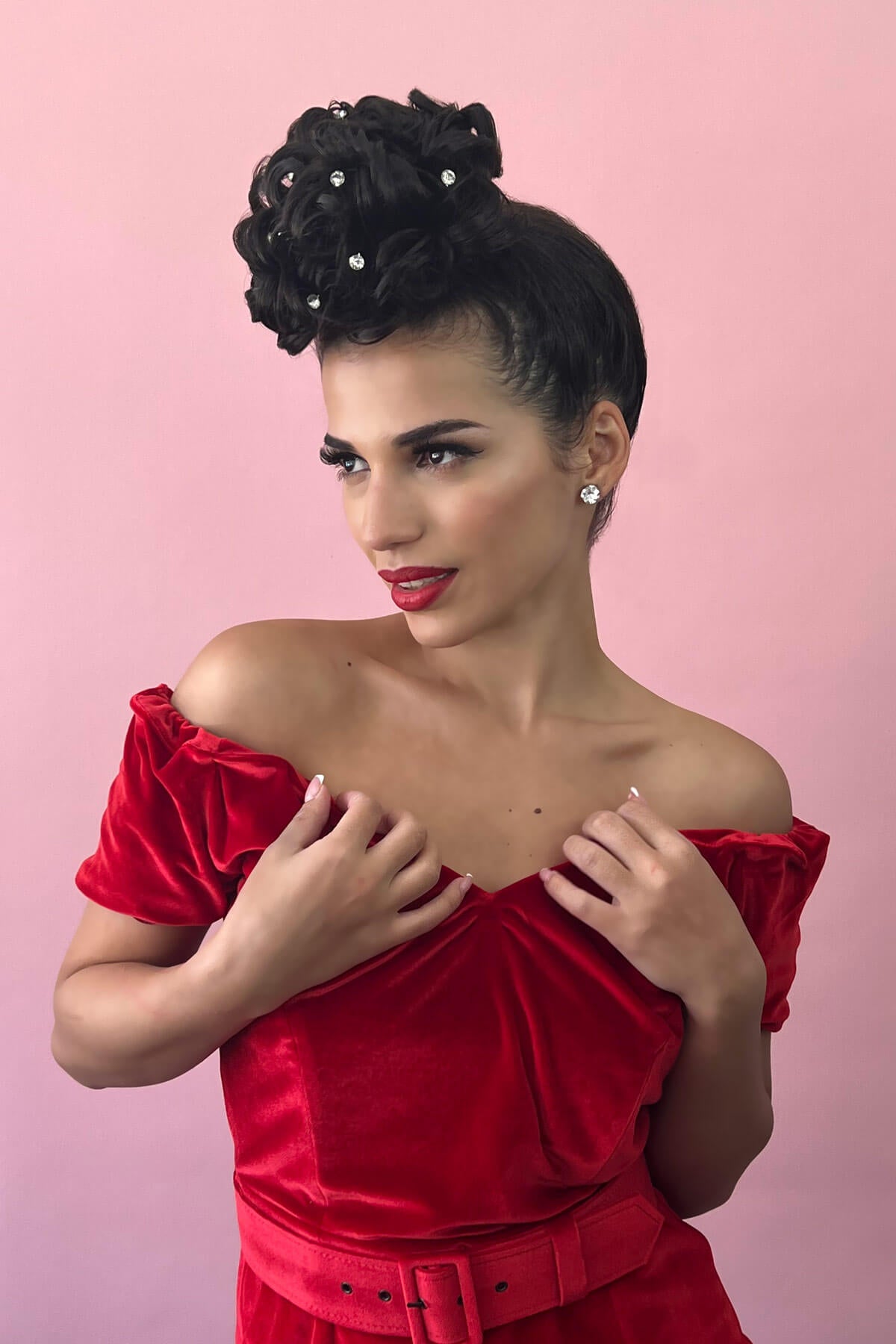 Woman wearing a red off-shoulder dress with a pink background. Hair poodle clip-in hairpiece bun vintage style 1950s: Lucille black 1B