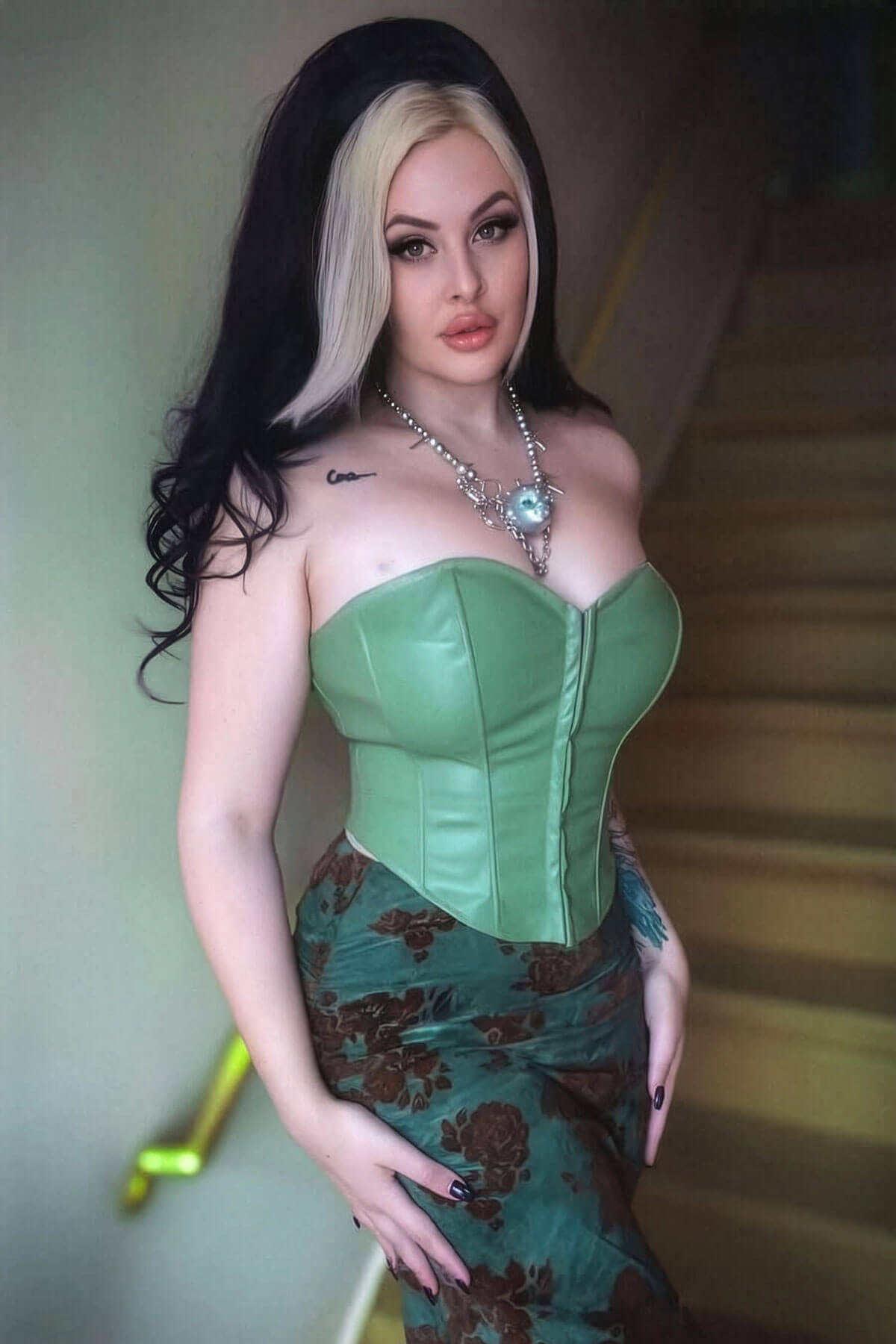 Woman wearing a green corset and floral dress against a neutral background. High crown long, wavy half wig and volume ring: Bardot jet black 1