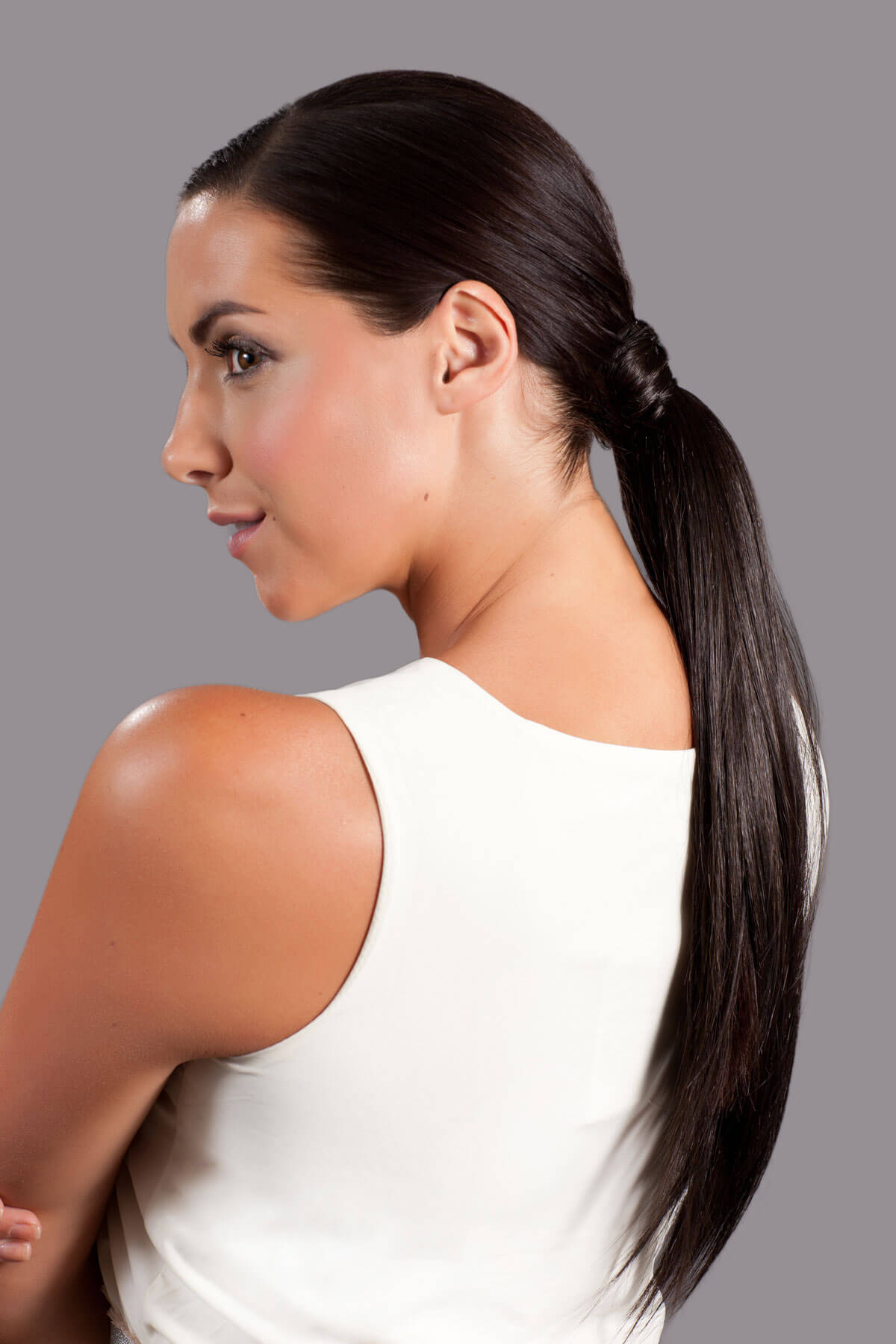 Woman with a high ponytail wearing a white sleeveless top on a gray background. Long sleek ponytail extension with gentle, layered waves: Mali dark brown 4