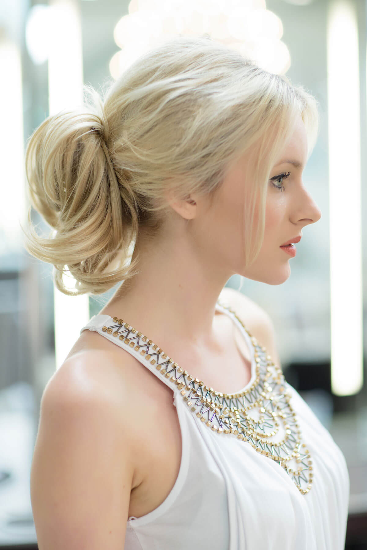 Woman with styled blonde hair wearing a white top with a decorative neckline. Clip in hairpiece, ponytail, short, flexihair: Leela honey blonde 24BH613