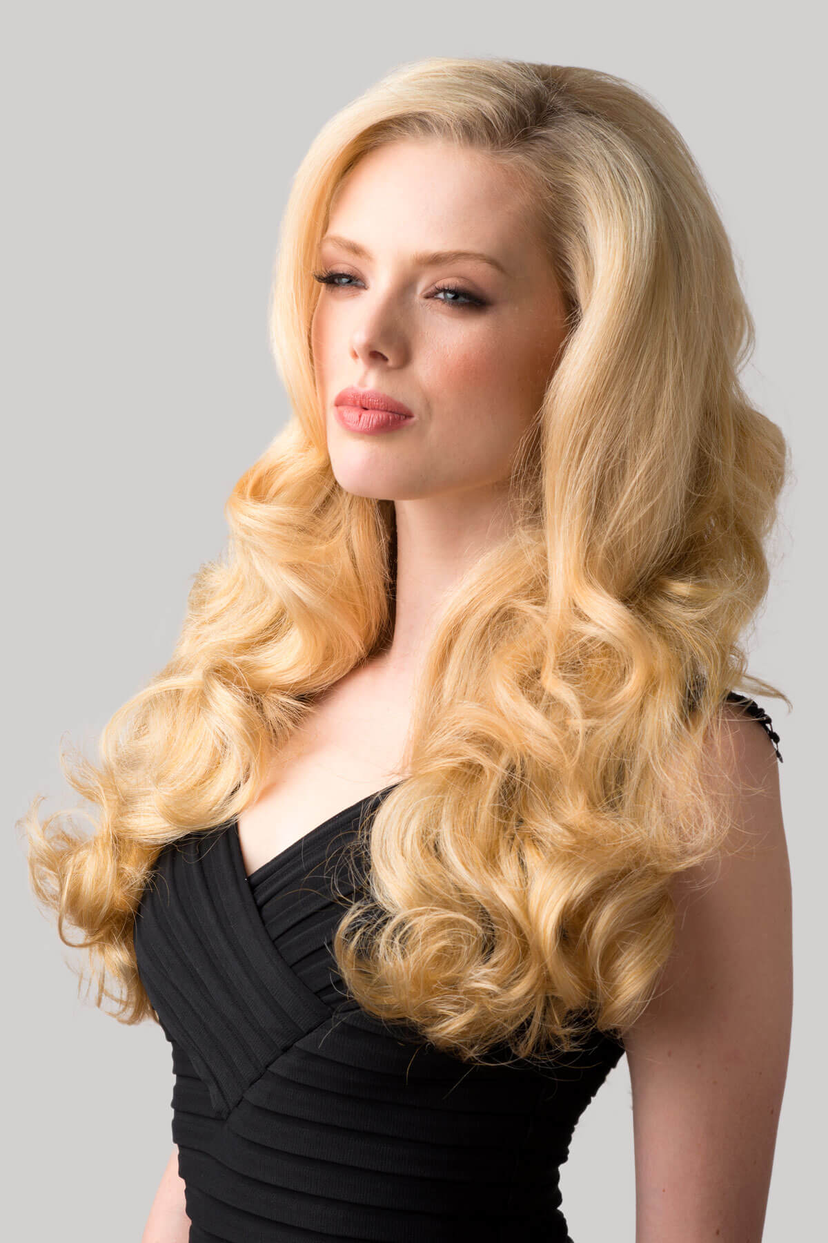 Woman with long, wavy blonde hair wearing a black dress against a gray background. Curly blonde half wig hairpiece extension (3/4 wig), long: Meana honey blonde 24BH613