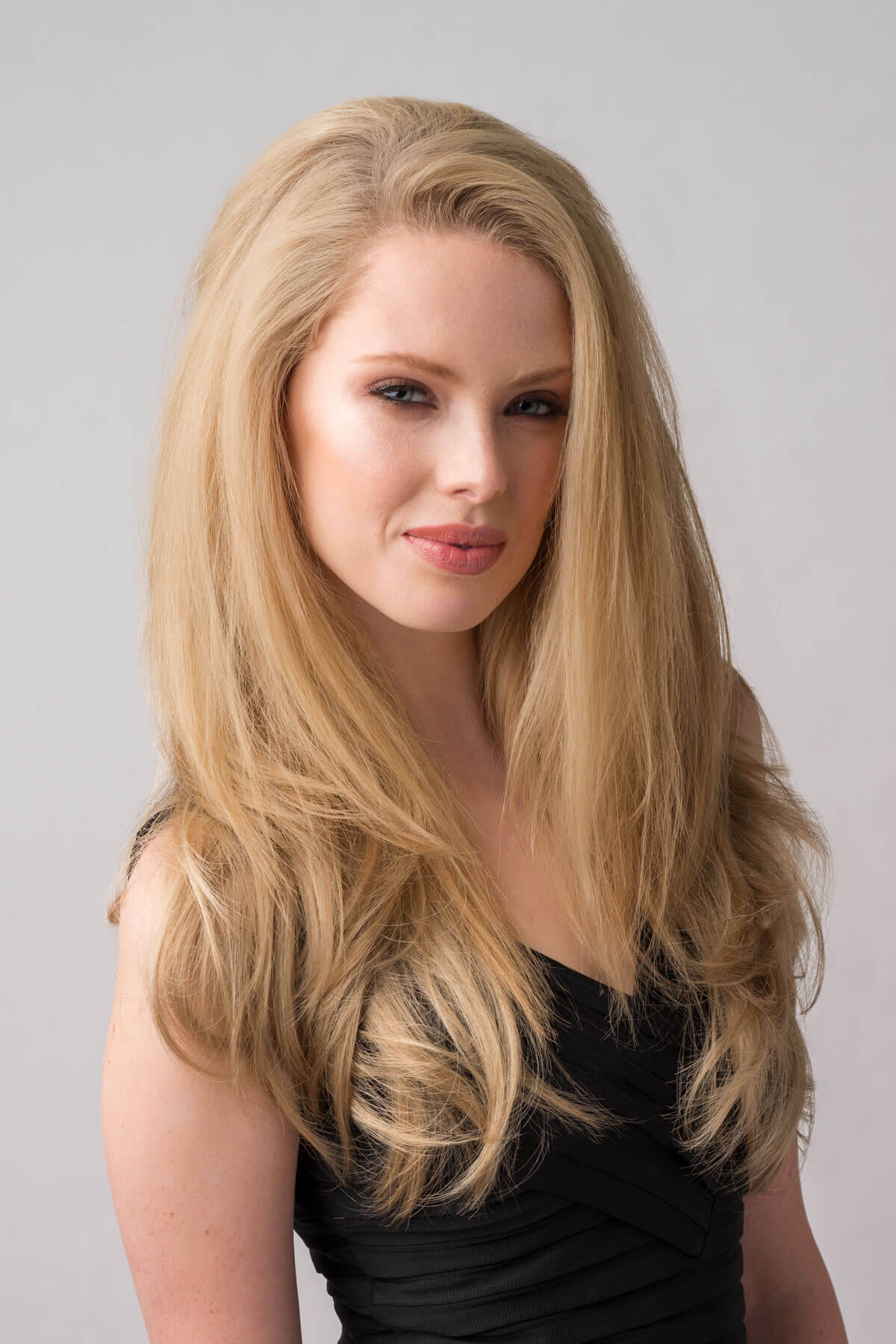 Woman with long blonde hair wearing a black top against a gray background. Straight blonde half wig hairpiece (3/4 wig), layered: Autumn blonde 24H613