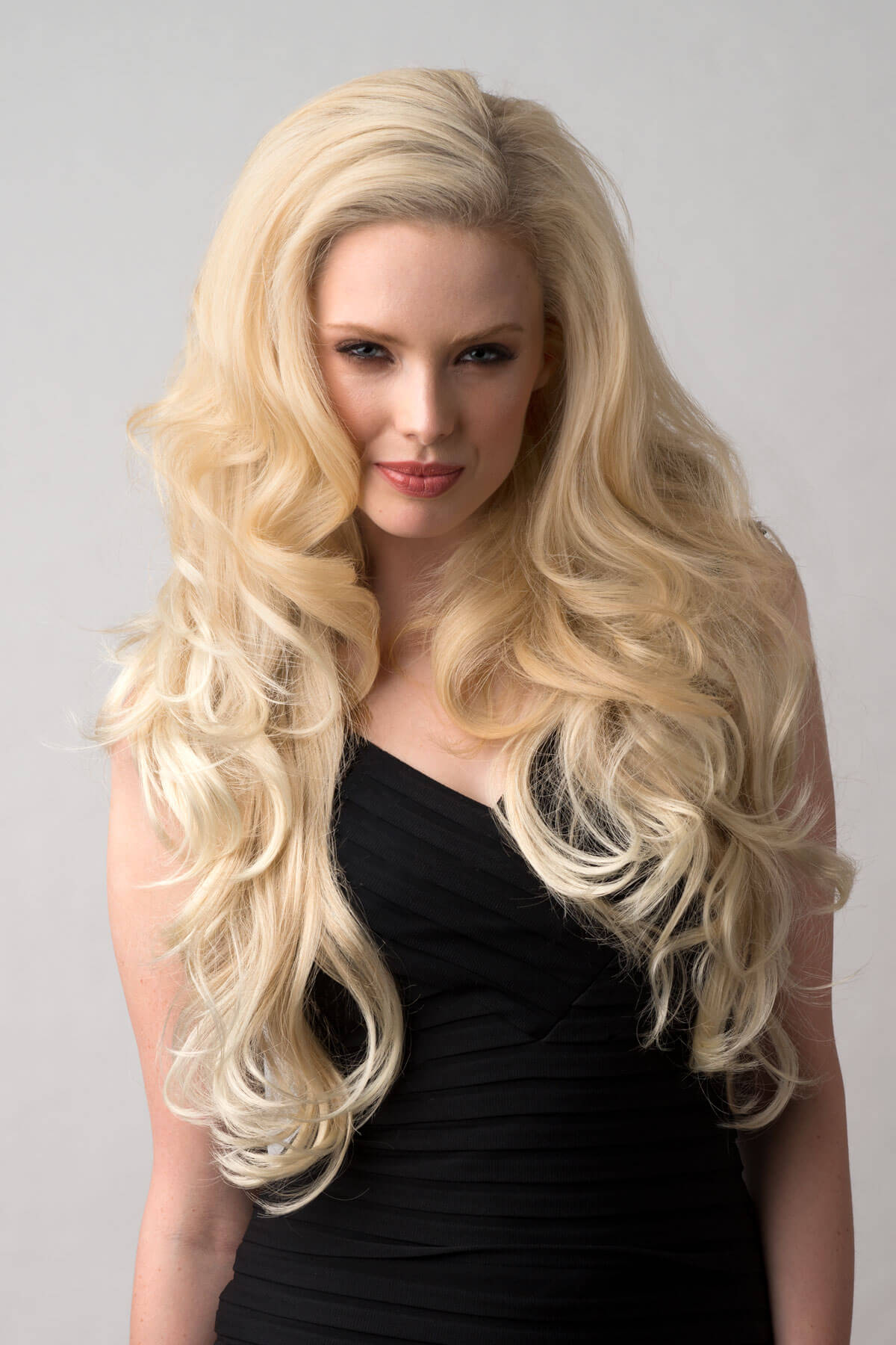 Woman with long blonde hair wearing a black top on a gray background. Long blonde 3/4 hairpiece (half wig), big loose curls: Zara blonde 613T24