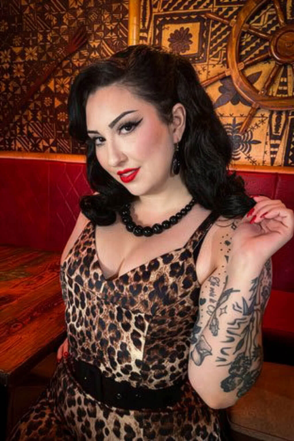 Woman in leopard print dress with tattoos sitting in a decorated room. 1950s style shoulder length half wig hairpiece with beautiful vintage waves: Onyx colour black 1B