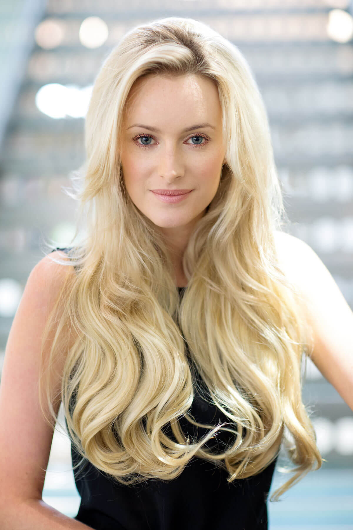 Woman with long blonde hair against a blurred indoor background. Heat styleable half wig hairpiece (3/4 wig), long wavy, Flexihair: Kate dark blonde 24BH613