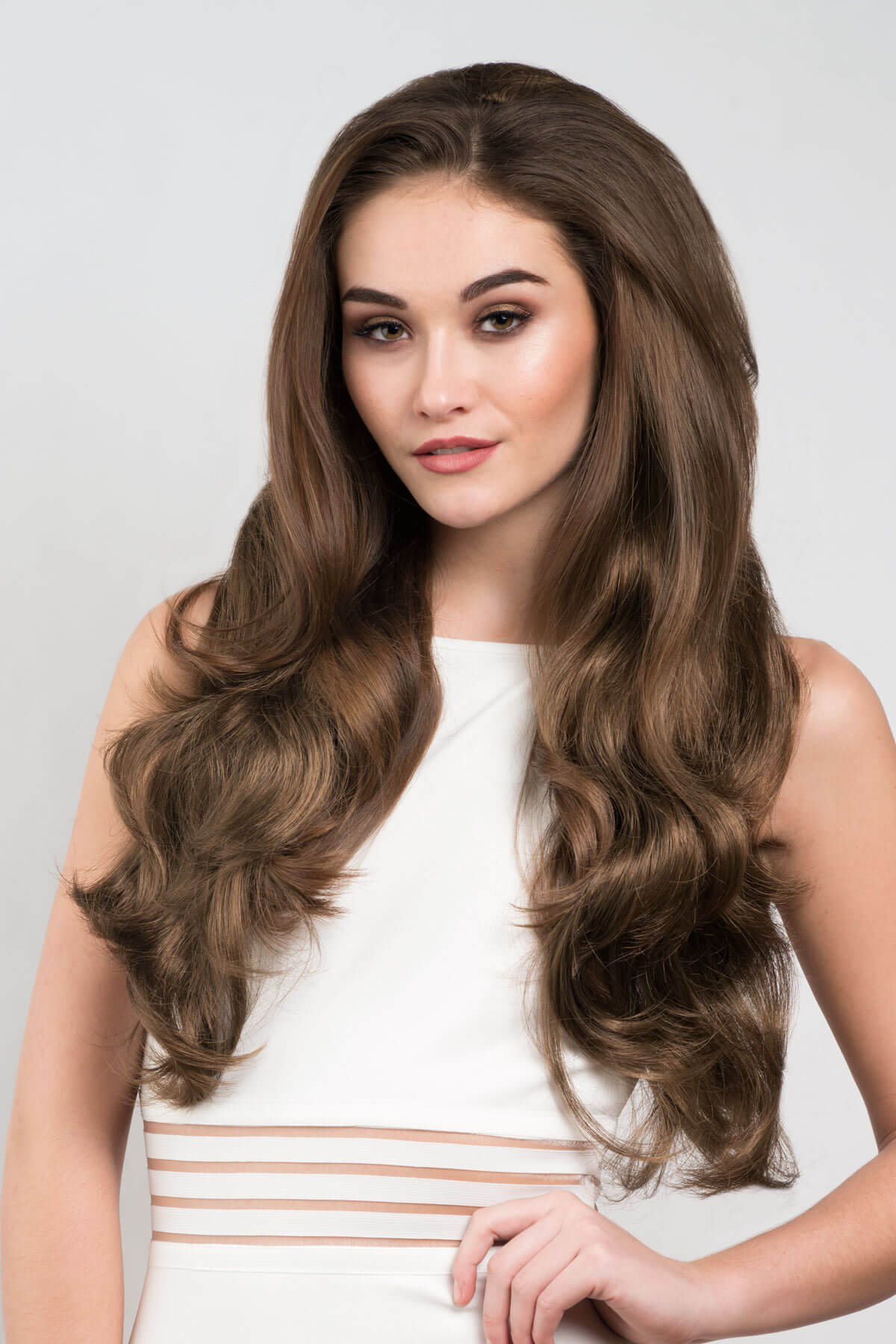 Woman with long, wavy brown hair wearing a white dress against a light gray background. Brown half wig hairpiece with long loose curls: Chelsea mocha brown 10