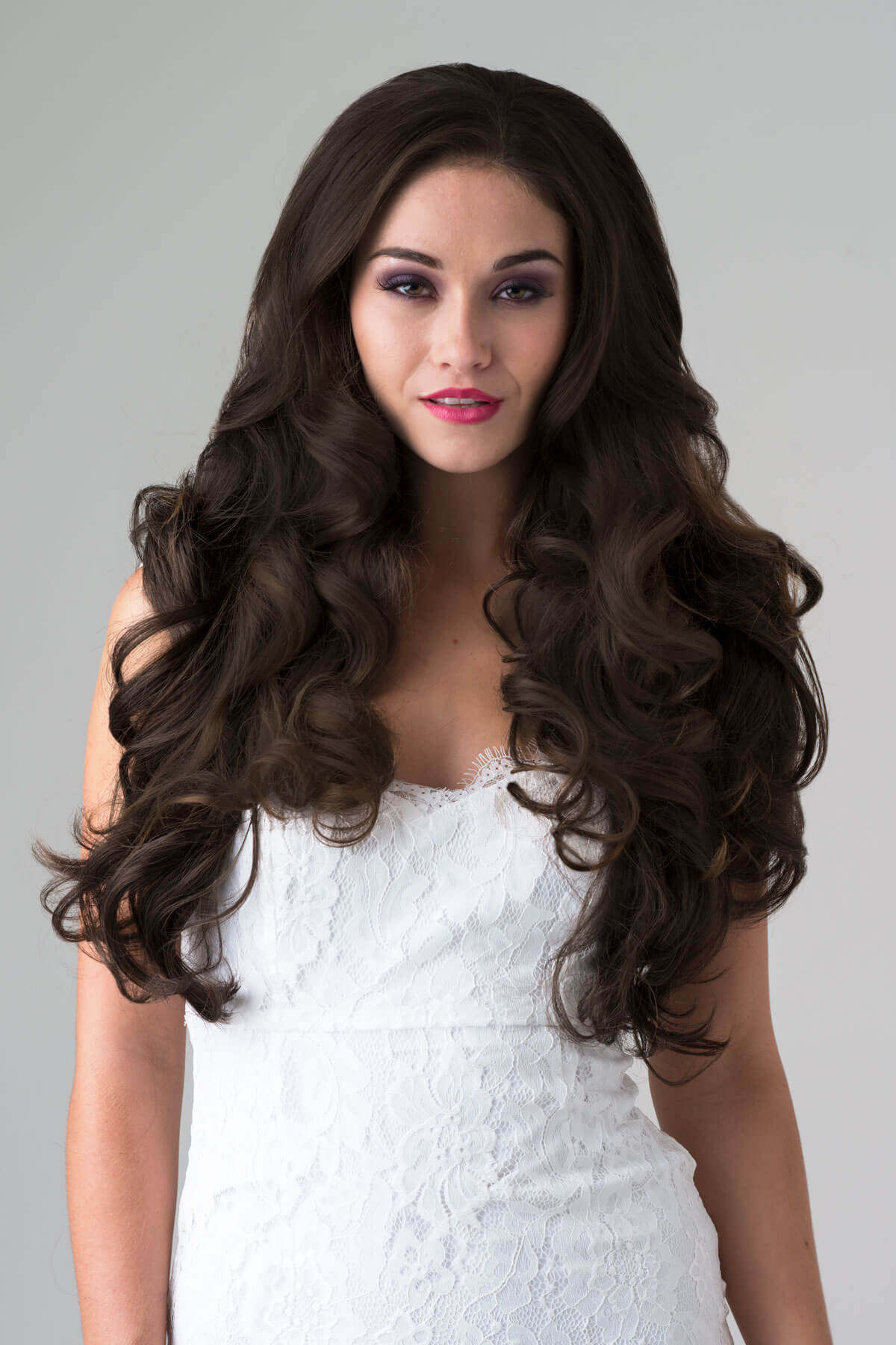 Woman with long, wavy brown hair wearing a white dress against a gray background. Brown half wig hairpiece with long loose curls: Chelsea dark chocolate brown 6