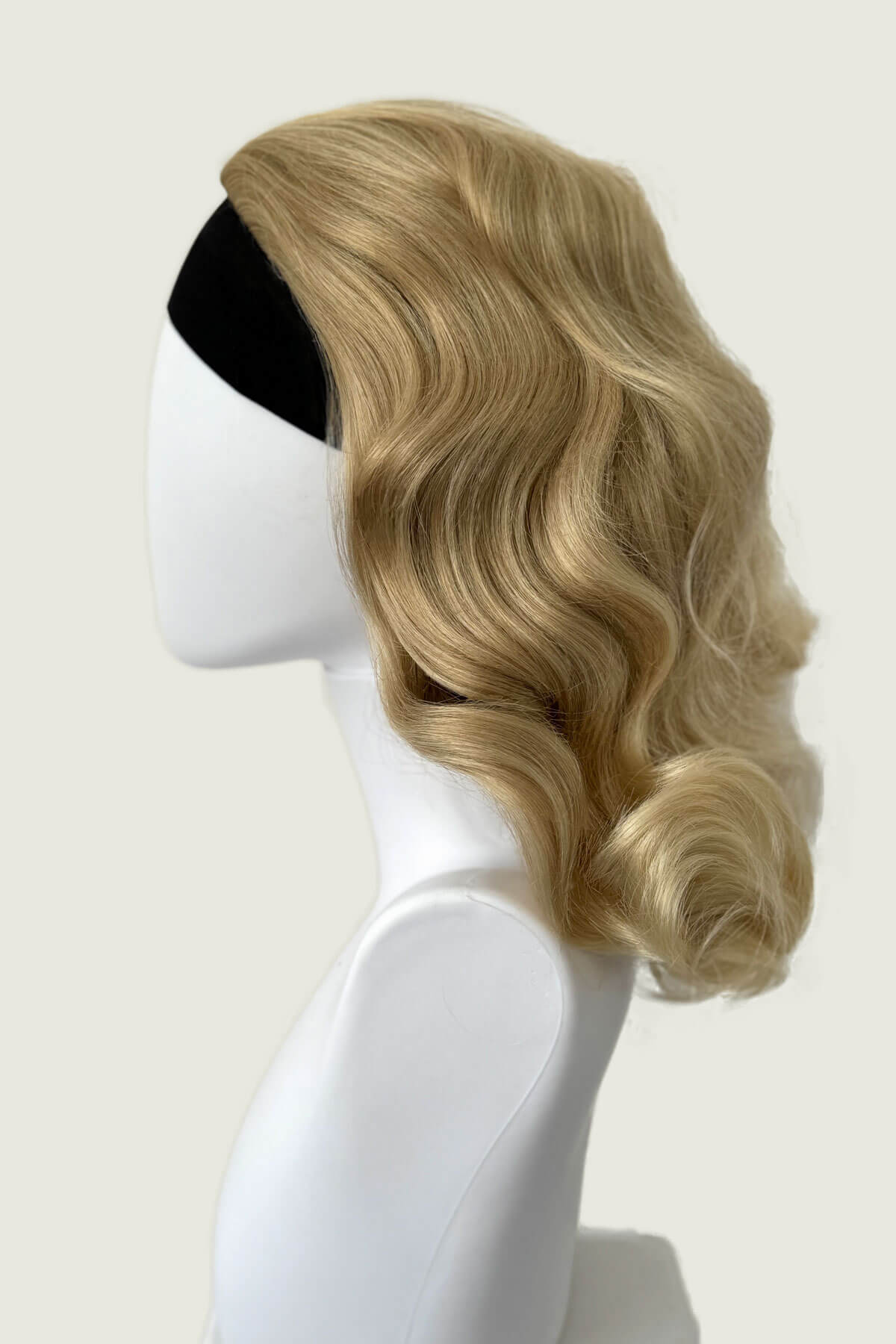 Blonde wig on a mannequin head against a white background, viewed from the side. 1950s style shoulder length half wig hairpiece with beautiful vintage waves: Onyx blonde 613T24