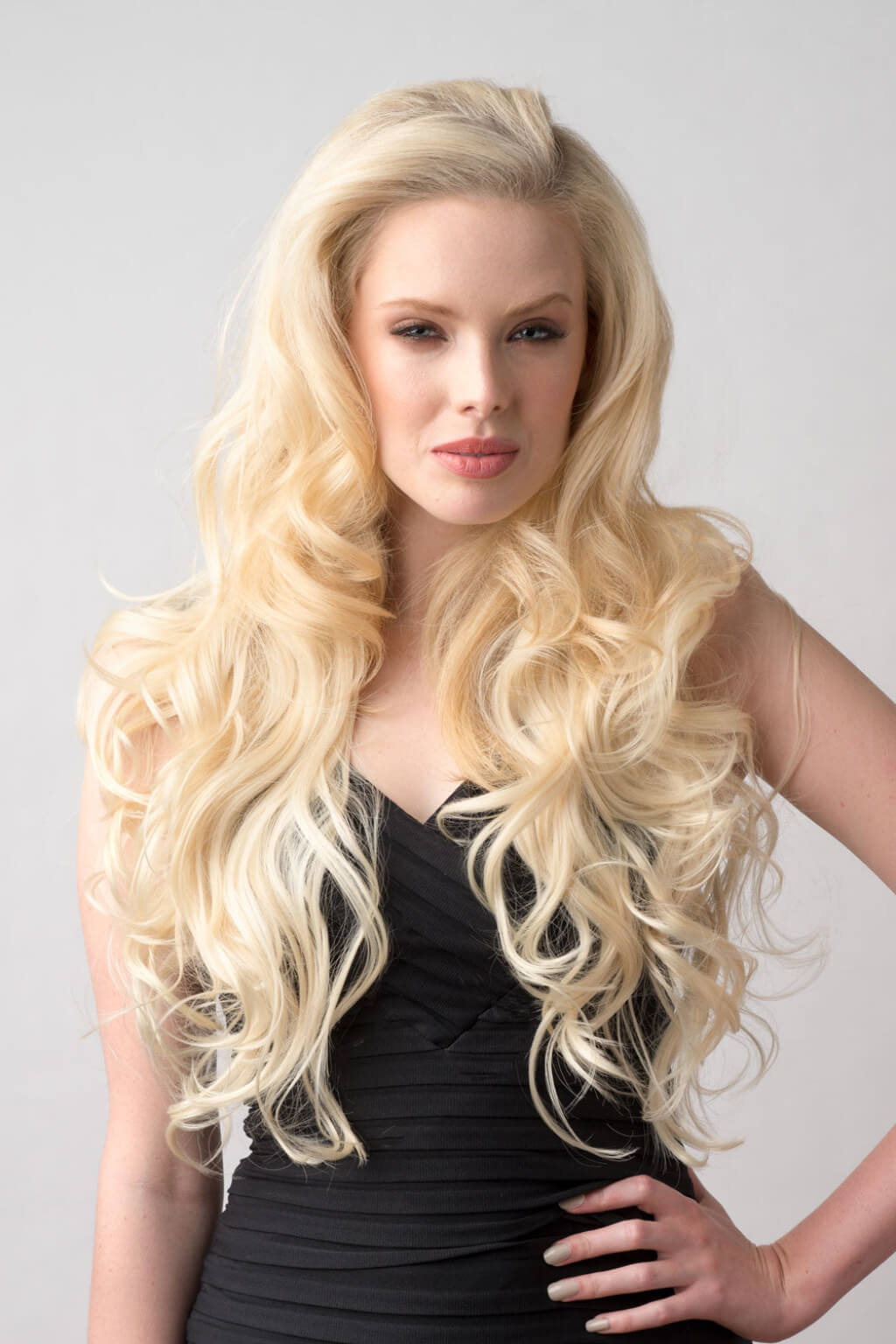 Woman with long, wavy blonde hair wearing a black top against a grey background. Blonde half wig hairpiece (3/4 wig), loose curls: Suki creamy blonde 613