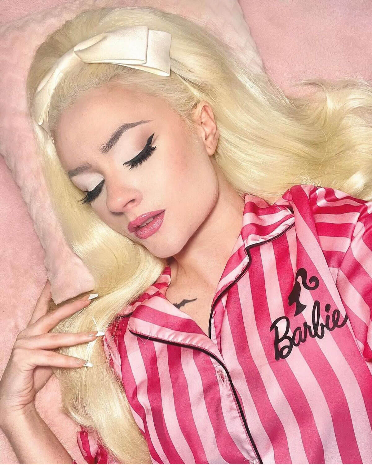 Person wearing pink and white striped pajamas with 'Barbie' logo on a pink background. High crown long, wavy half wig and volume ring: Bardot blonde 613
