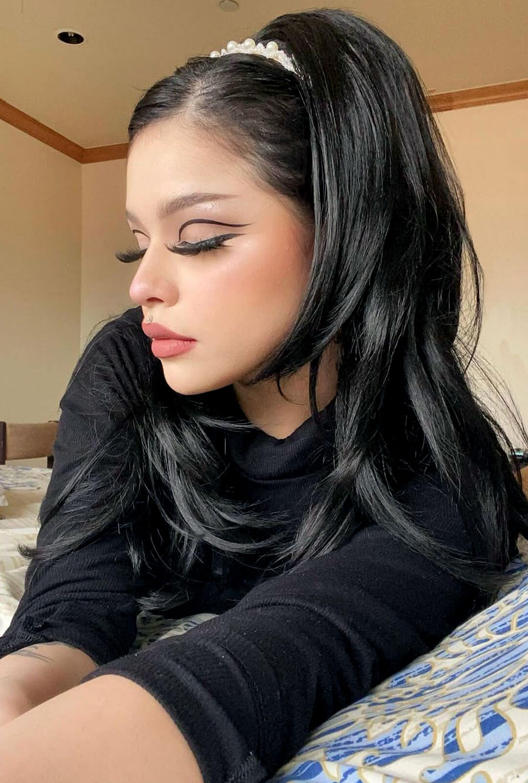 Woman with long black hair wearing a headband, sitting indoors. 1960s style hair, high crown long straight and layered half wig: Juliette black 1B