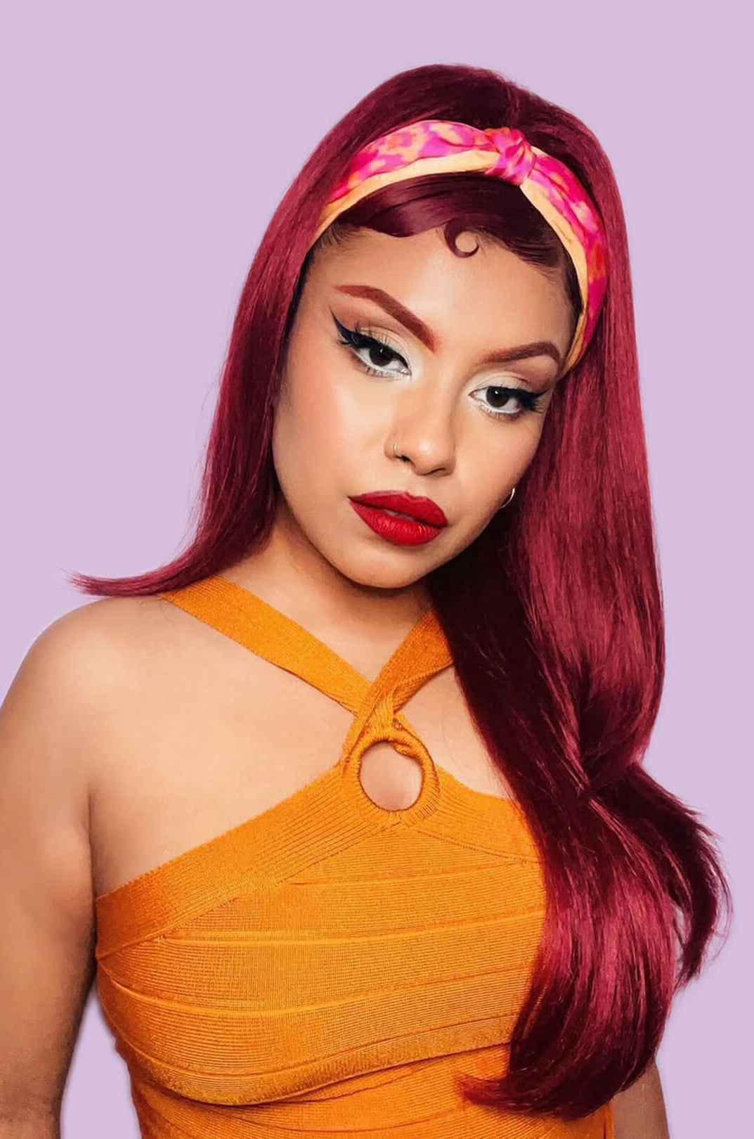 Woman with vibrant red hair and orange dress against a light purple background. 1960s style hair, high crown long straight and layered half wig: Juliette wine red 39