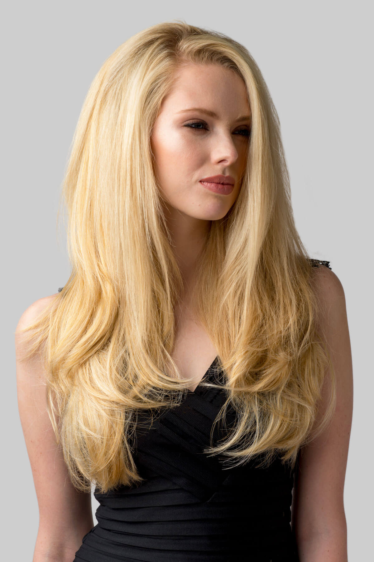 Woman with long blonde hair wearing a black dress against a gray background. Straight blonde half wig hairpiece (3/4 wig), layered: Autumn honey blonde 24BH613