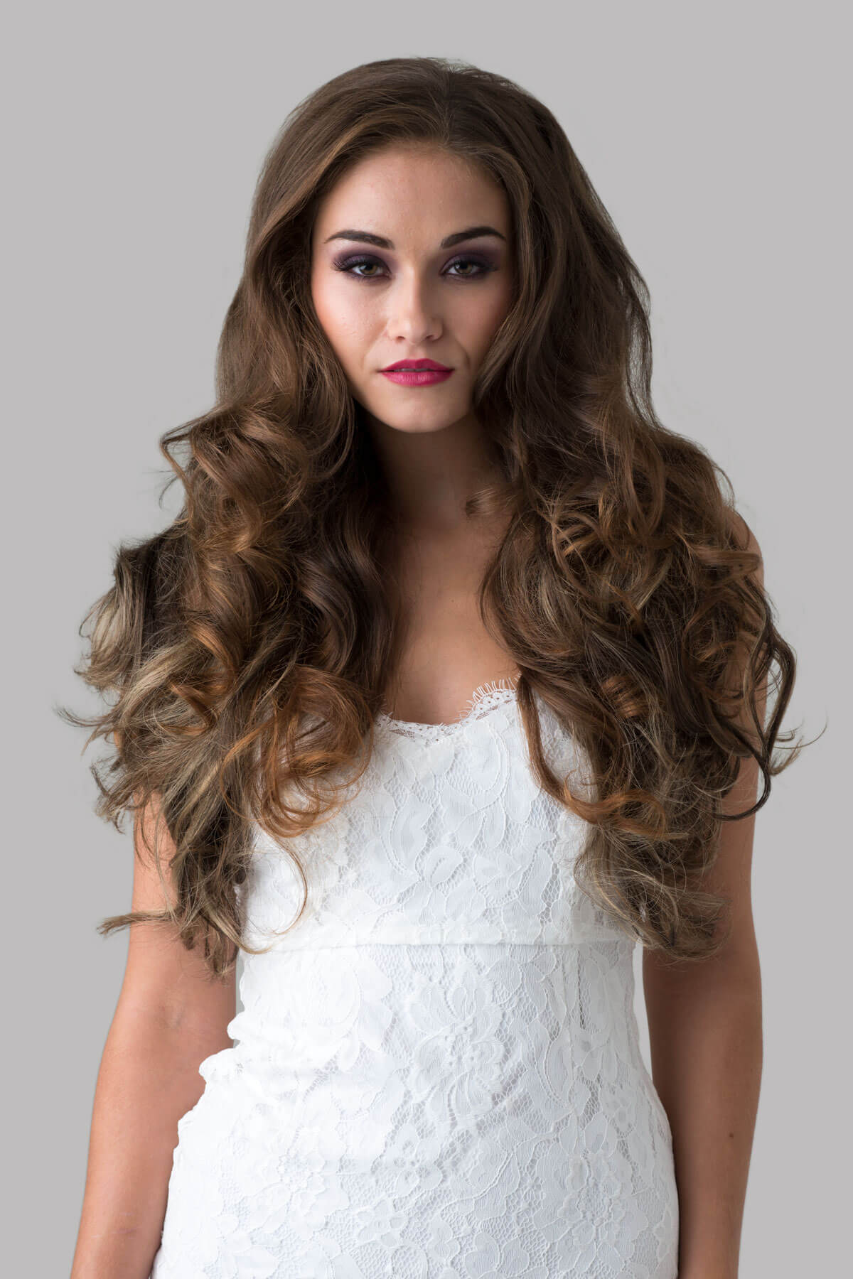 Woman with long, wavy hair wearing a white lace dress against a gray background. Brown half wig with blonde highlights with big loose curls: Nickie