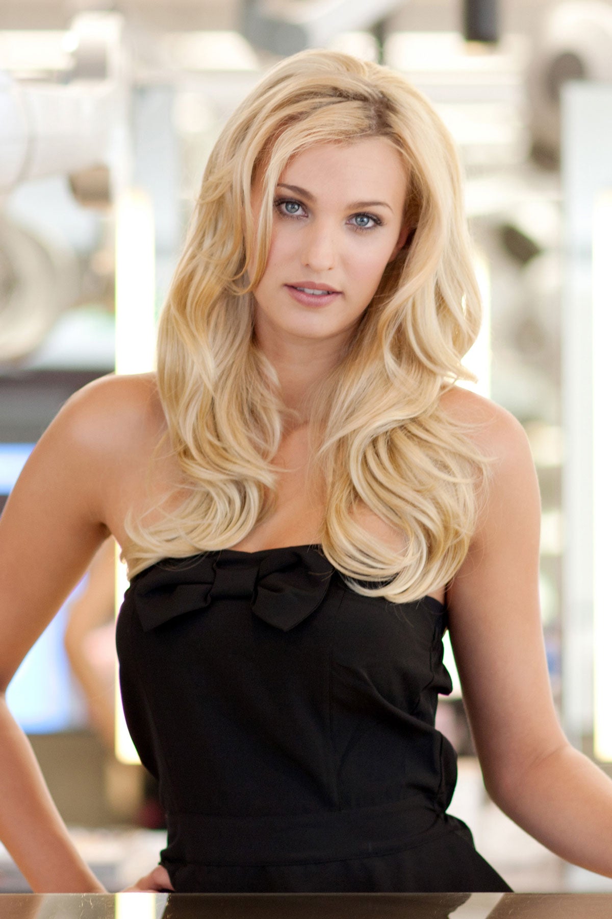 Woman with long blonde hair wearing a black dress in an indoor setting. Heat styleable half wig hairpiece (3/4 wig), long, wavy, Flexihair: Michela blonde 24BH613