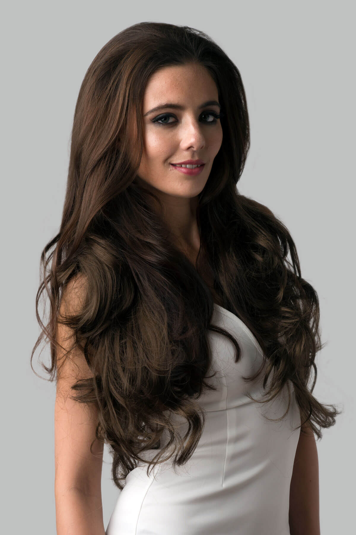 Woman with long, wavy brown hair wearing a white dress against a gray background. Brown half wig hairpiece, full, long with gentle waves: Taya mocha brown 10