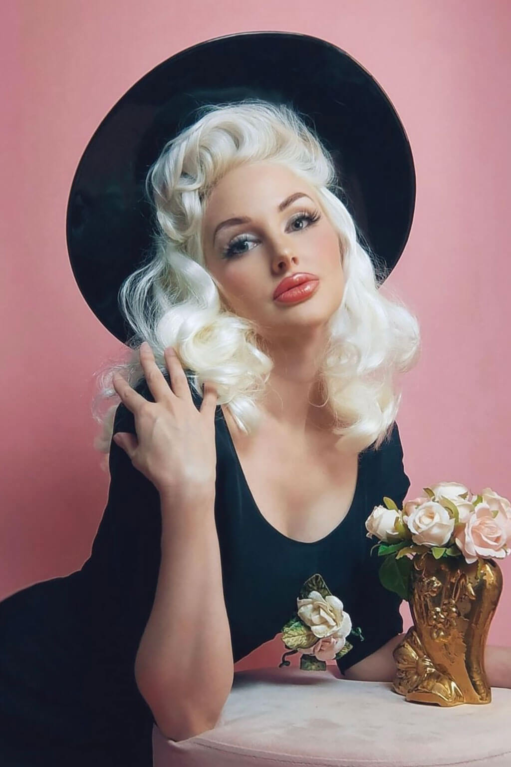 A person with shoulder-length black waves wearing a black sleeveless dress against a pink background. 1950s style shoulder length half wig hairpiece with beautiful vintage waves: Onyx platinum blonde 1001