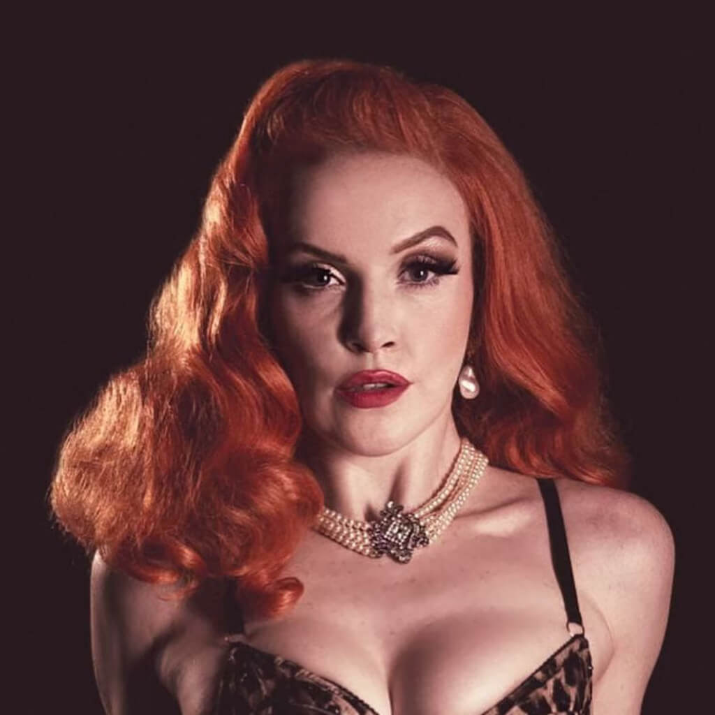Woman with red hair and vintage makeup wearing a necklace against a dark background. 1950s style shoulder length half wig hairpiece with beautiful vintage waves: Onyx ginger