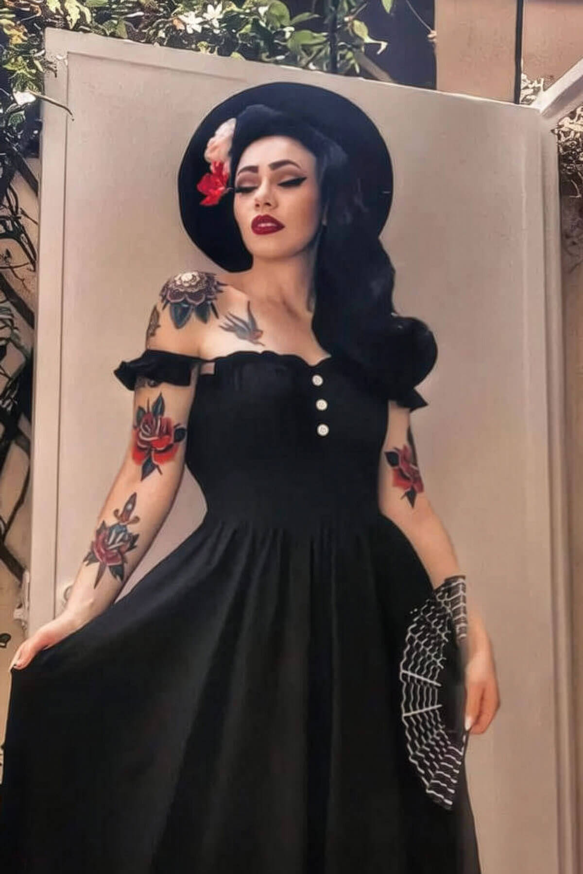 Woman in a black dress with tattoos and a hat, standing against a neutral background. 1960s Retro high crown, half wig hairpiece, long with hair flip: Priscilla jet black 1