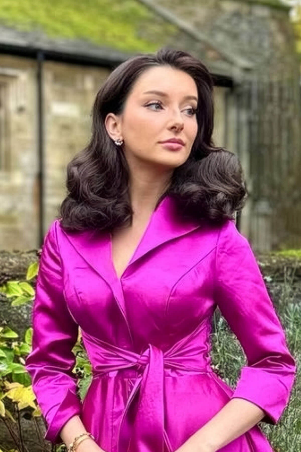 Woman in a bright pink dress standing outdoors with greenery in the background. 1940s style half wig hairpiece with beautiful marcel waves: Virginia chestnut brown 8