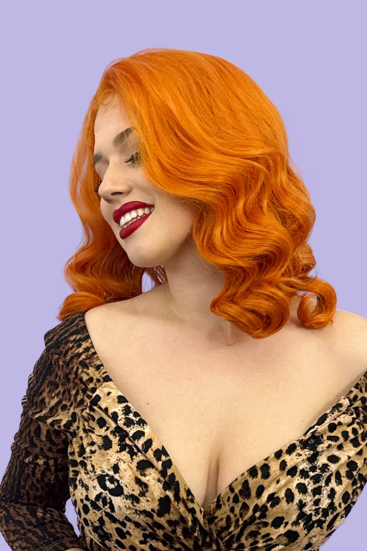 Woman with vibrant orange hair and red lipstick wearing a leopard print dress against a purple background. Ginger pinup lace front wig, Dita Von Teese style vintage style wig with finger waves Lula