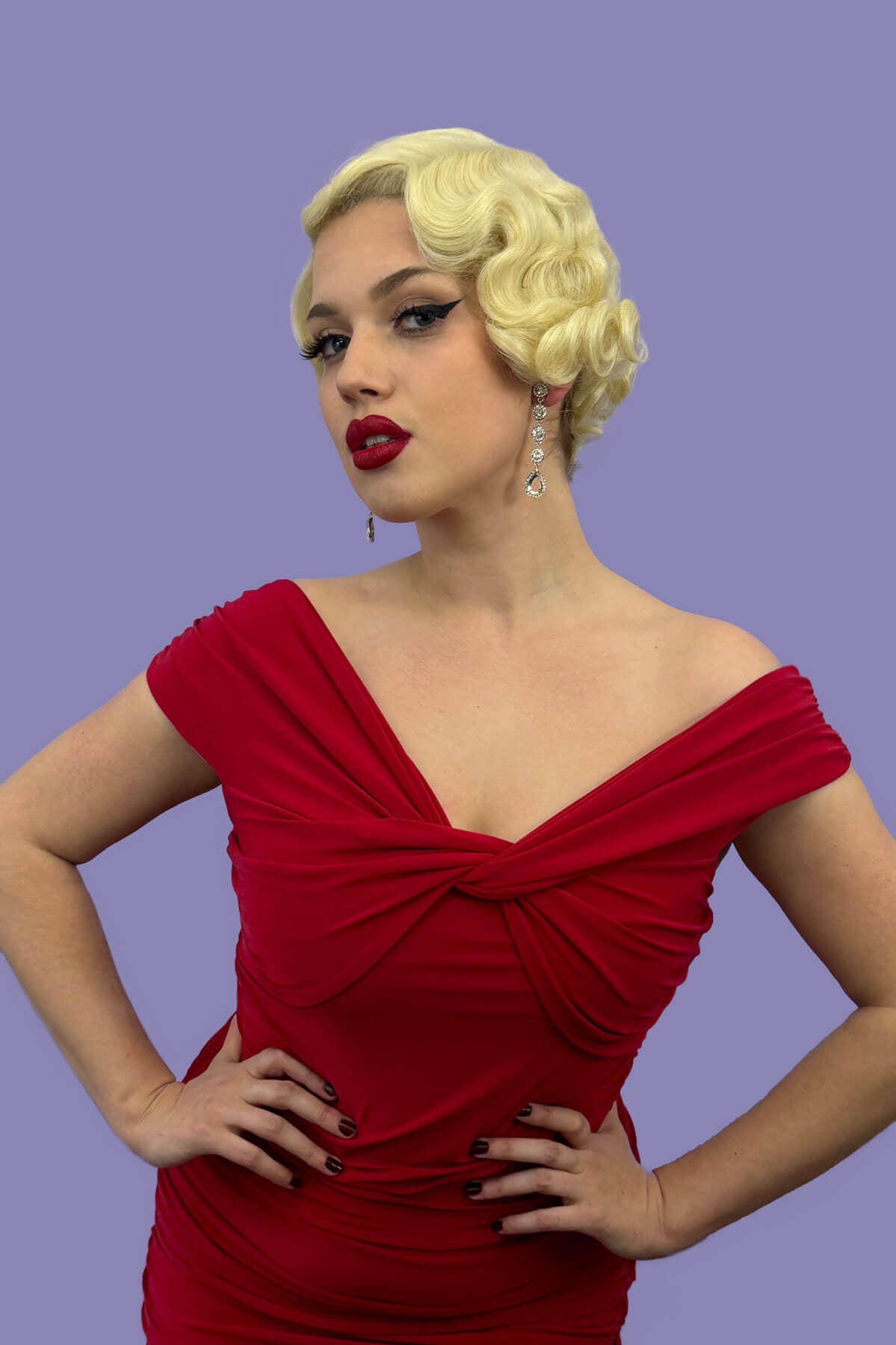 A person wearing a blonde vintage style lace front wig with finger waves, posing in a red dress against a purple background. Blonde pinup wig, lace front, vintage style: Odette champagne blonde 613