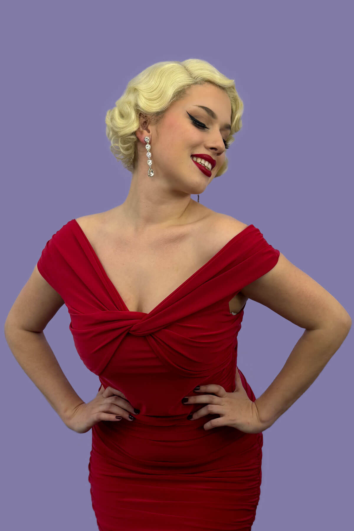 A person wearing a blonde vintage style lace front wig with finger waves, posing in a red dress against a purple background. Blonde pinup wig, lace front, vintage style: Odette champagne blonde 613
