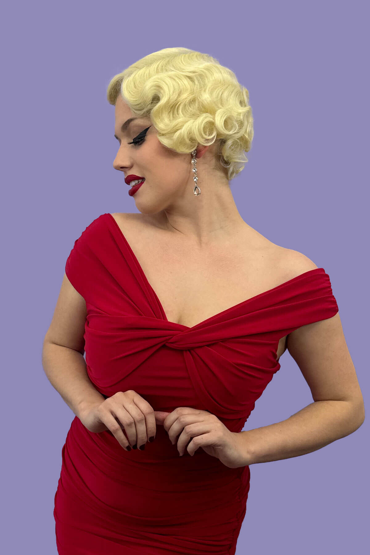 A person wearing a blonde vintage style lace front wig with finger waves, posing in a red dress against a purple background. Blonde pinup wig, lace front, vintage style: Odette champagne blonde 613