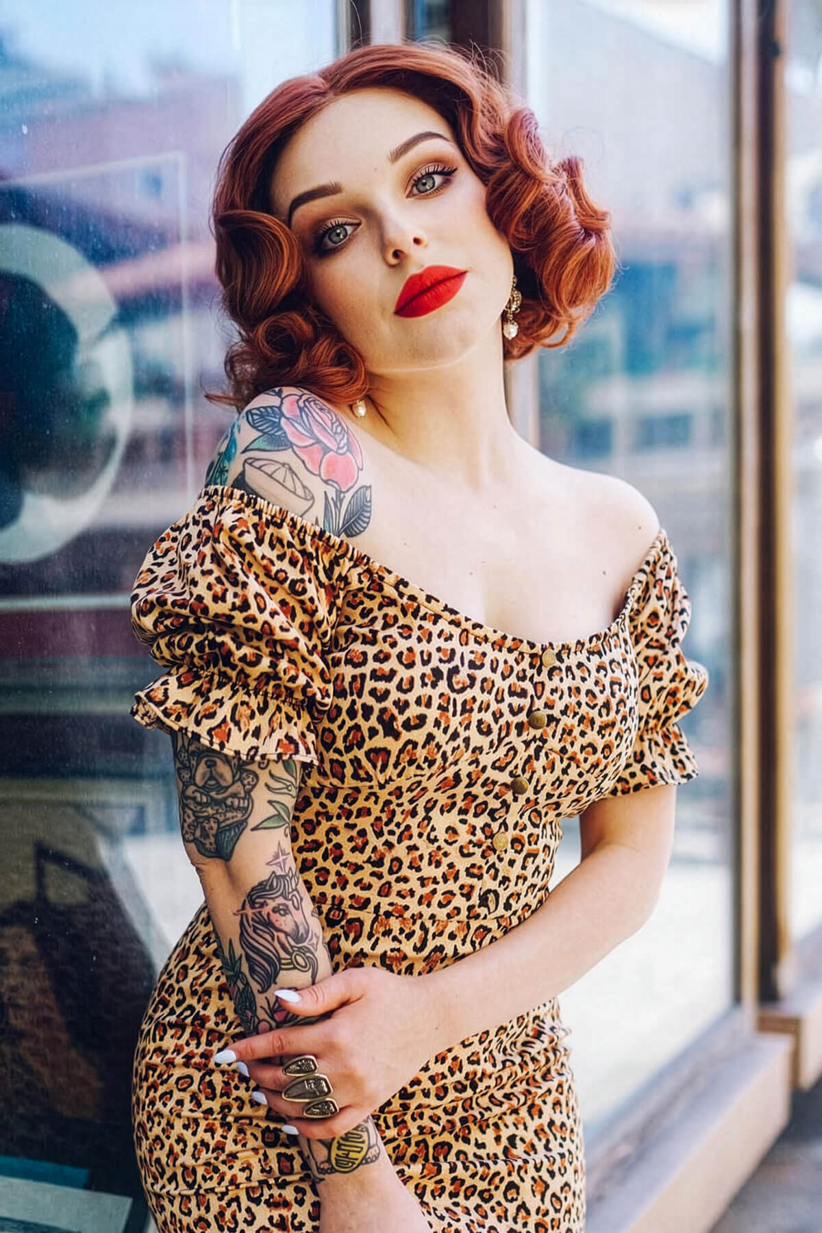 Woman wearing a leopard print dress with tattoos and red lipstick, standing in front of a glass window. Red, auburn & blonde pinup wig, lace front, vintage style: Ginny