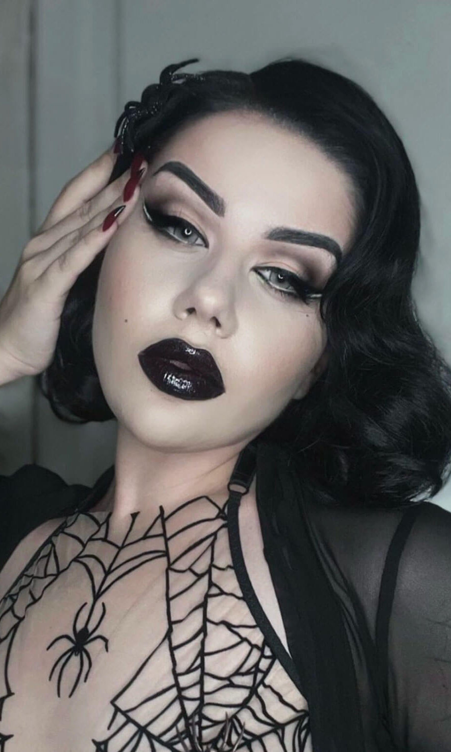 Woman with dark makeup and spider-themed sheer top against a neutral background. Black lacefront wig, pinup/vintage style, mid length with finger waves: Lilith