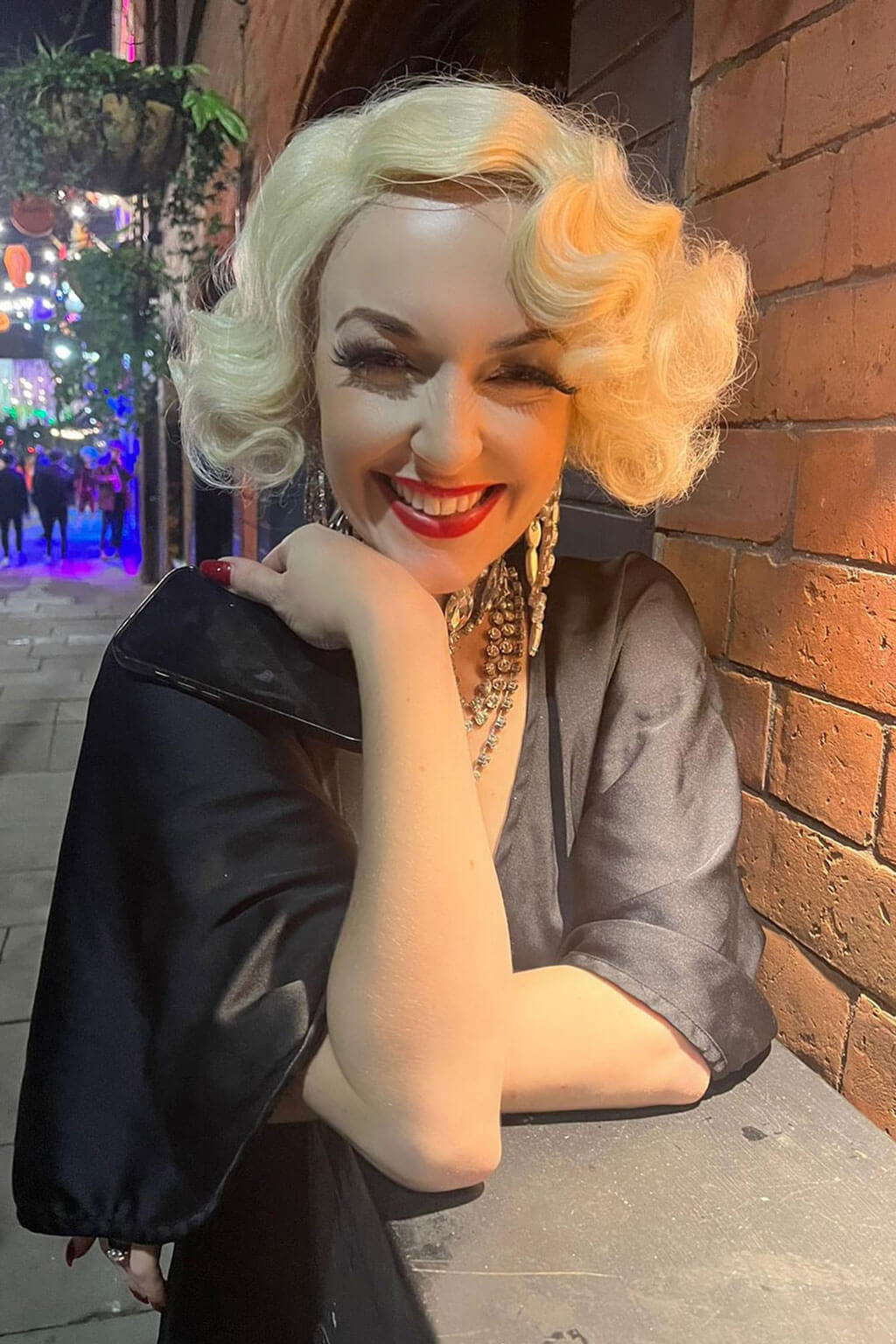 Mannequin with blonde hair and red lipstick against a brick wall at night. Blonde pinup wig, lace front, vintage style: Odette champagne blonde 613