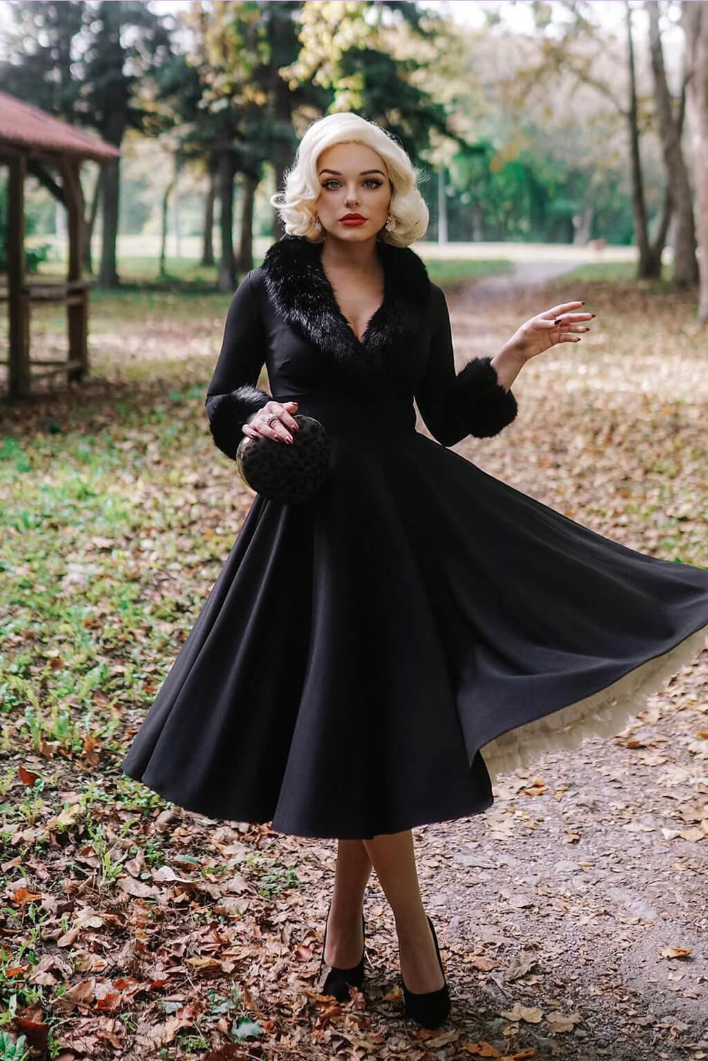 Woman in a black coat with fur trim standing in a park with fallen leaves. Blonde pinup wig, lace front, vintage style: Marilyn white blonde 1001