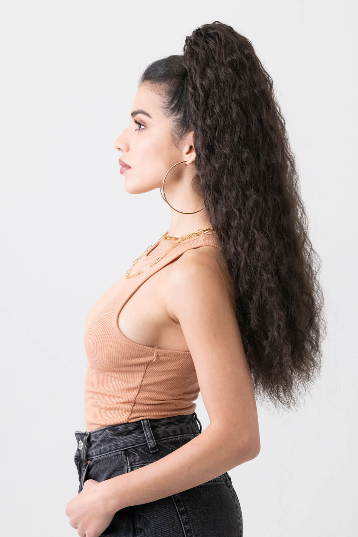 Woman wearing a beige tank top and black pants with gold jewelry on a white background. Extra long ponytail extension with long loose spirals: Kaia dark brown 4