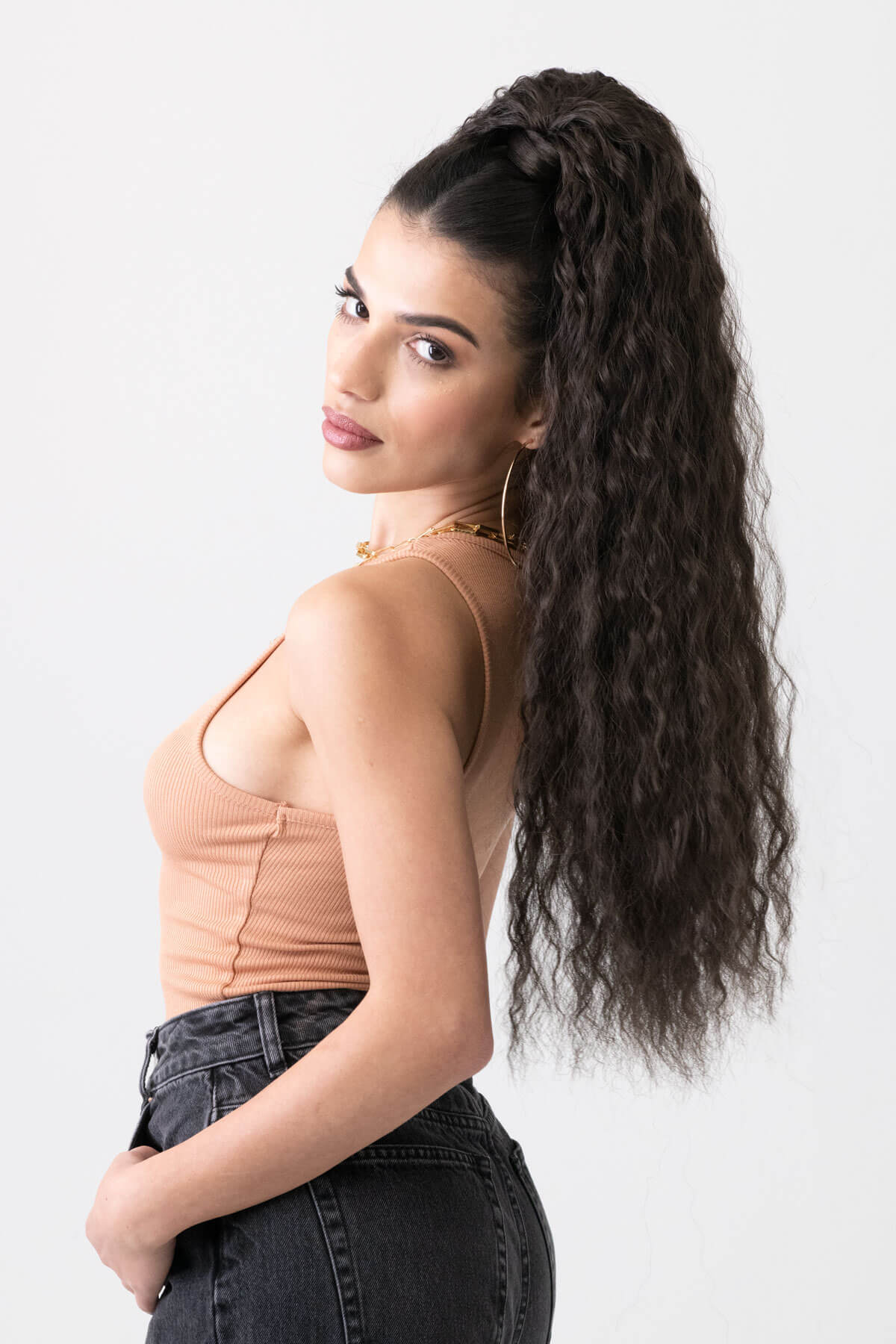 Woman wearing a beige tank top and black pants with gold jewelry on a white background. Extra long ponytail extension with long loose spirals: Kaia dark brown 4