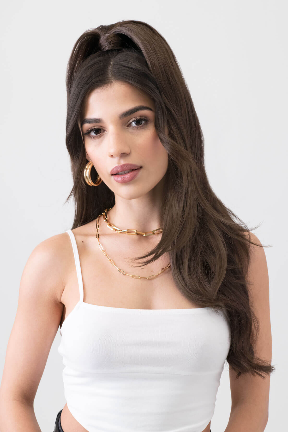 Woman wearing a white top and gold jewelry against a plain background. Long sleek ponytail extension with gentle, layered waves: Mali chestnut brown 8