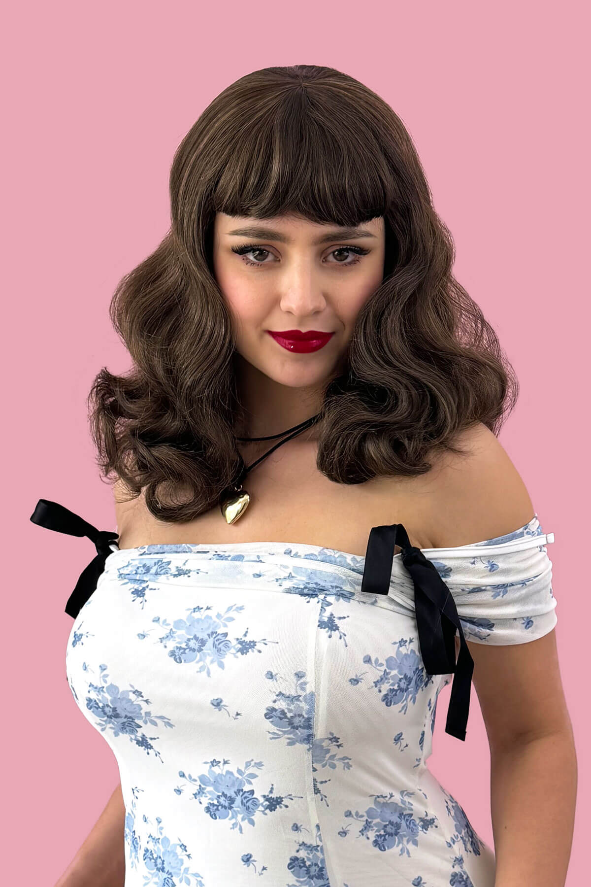 Woman wearing a floral off-shoulder top with black bows against a pink background. Brown pinup style wig, curled with short fringe: Carolina Brown and blonde 8H27