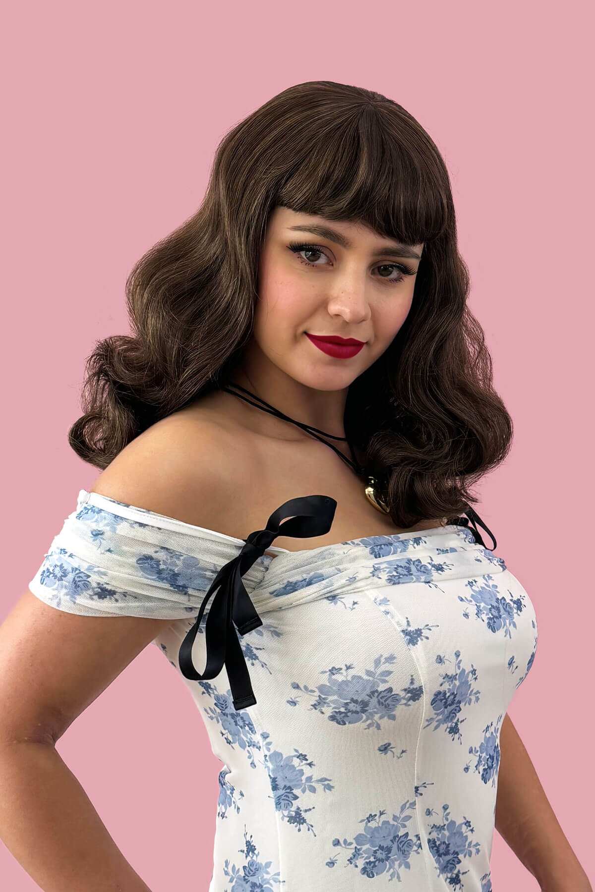 Woman wearing a floral off-shoulder top with a black bow against a pink background. Brown pinup style wig, curled with short fringe: Carolina Brown and blonde 8H27