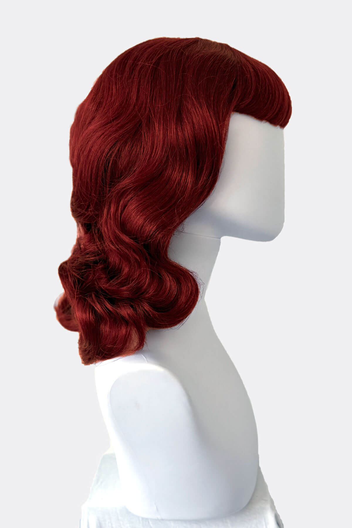 Red wig from the side on a mannequin head against a white background. Copper red 1950s pinup wig, curled with short fringe: Kitty